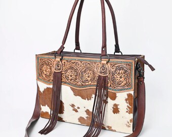 May include: A brown leather tote bag with a white and brown cowhide pattern. The bag has a top zipper closure, two top handles, and a detachable shoulder strap. The front of the bag features intricate tooled leather designs with turquoise accents and fringe.