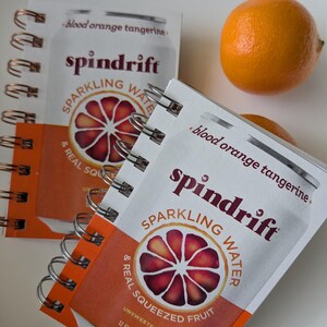 May include: Two spiral-bound notebooks featuring a graphic of a Spindrift blood orange tangerine sparkling water can. The cover displays the brand name, a citrus slice illustration, and the text 12 FL OZ (355ML). A whole orange sits nearby.