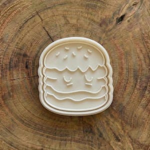 Puede incluir: Cortador de galletas de plástico blanco con forma de hamburguesa con una cara sonriente. El cortador tiene un pan, una hamburguesa y otros ingredientes. El cortador está sobre una superficie de madera.