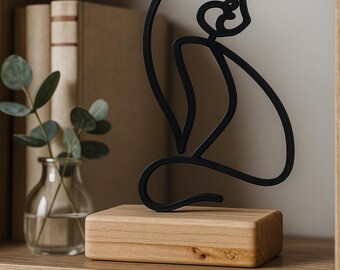 Op de afbeelding: Een zwarte metalen katsculptuur in minimalistische stijl staat op een natuurlijke houten blok. De sculptuur staat op een houten plank, met links een kleine glazen vaas met groen. Op de achtergrond zijn boeken te zien.