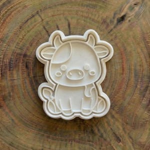 Puede incluir: Cortador de galletas blanco con forma de vaca. El cortador presenta un diseño de vaca de dibujos animados con un cuerpo redondeado, cuernos y una mancha en la cabeza. El cortador está sobre una superficie de madera.