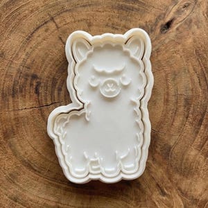 May include: A white plastic llama-shaped cookie cutter. The cutter has a detailed outline of the animal, including its ears, face, and body. The cutter is designed for making cookies or other treats in the shape of a llama.