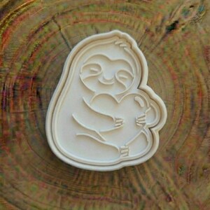 May include: A white plastic cookie cutter in the shape of a smiling sloth holding a heart. The sloth has a simple, cartoonish design with curved lines. The cutter is set against a wood grain background.