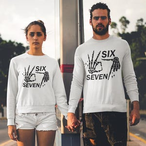 May include: Two people wearing matching white sweatshirts with the text "SIX SEVEN" and a skeleton hand design. The person on the left is wearing white shorts and the person on the right is wearing black shorts. They are holding hands.