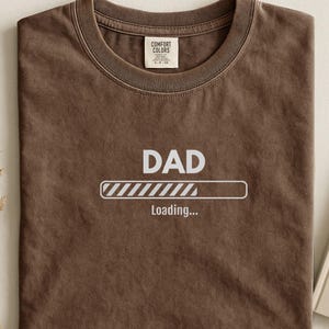 May include: Brown t-shirt with the word "DAD" above a loading bar graphic. The phrase "Loading..." is below the bar. The shirt is folded and has a tag in the upper center.