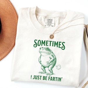 May include: Cream-colored t-shirt with a green frog graphic and the text "SOMETIMES I JUST BE FARTIN'". The shirt has a round neckline and short sleeves. A gold necklace with a pendant is draped on the shirt.