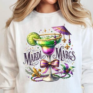 May include: White long-sleeve sweatshirt featuring a colorful margarita design with the text "Mardi Margs." The margarita glass is green and purple, with a lime slice and a small umbrella. The design includes beads and a bow.