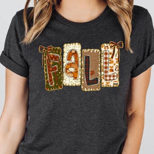 May include: A dark charcoal gray short-sleeve t-shirt featuring the word FALL in large, block-style letters. Each letter has a unique pattern, including plaid, polka dots, and woodland motifs, accented with small brown bows at the top corners.