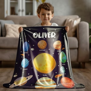 May include: A dark blue blanket featuring a solar system design with planets and stars. The blanket has the name "OLIVER" in white letters. Planets include Mercury, Venus, Earth, and Saturn. The blanket is held by a young child.