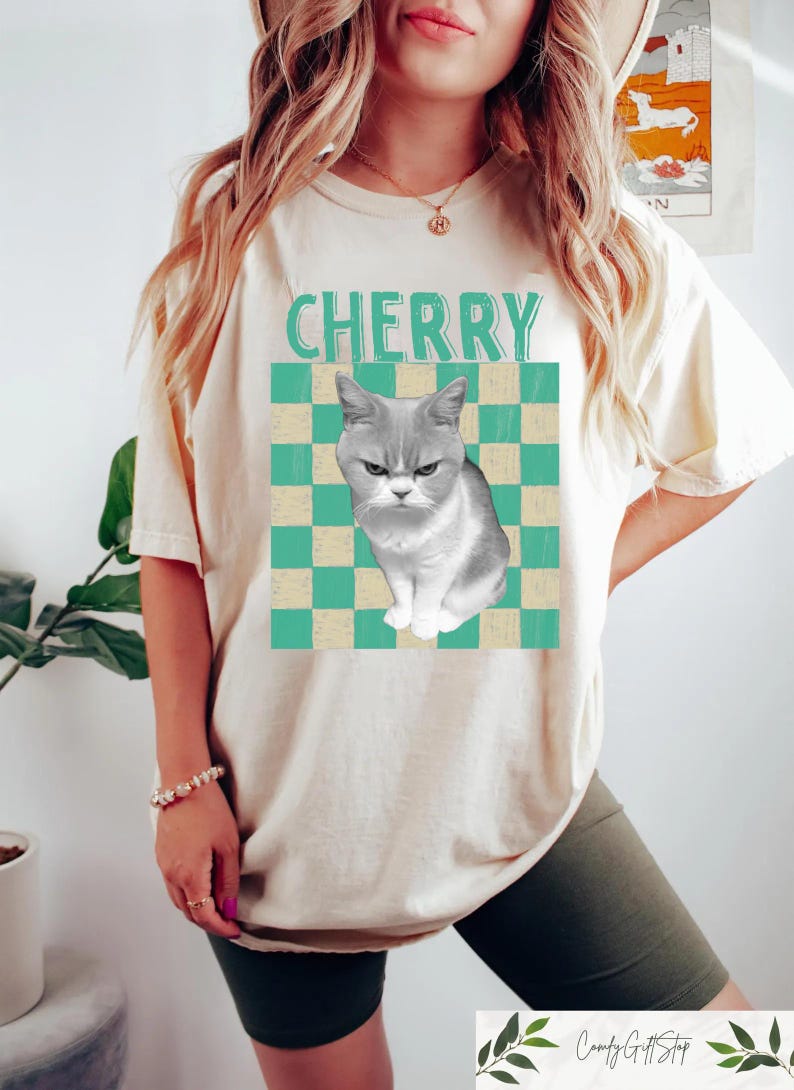 May include: Cream-colored t-shirt featuring a grumpy gray cat graphic on a teal and cream checkered background. The word "CHERRY" is printed above the cat. The shirt has short sleeves and a relaxed fit.