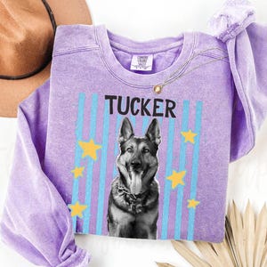 May include: A lilac-colored sweatshirt featuring the name "TUCKER" above a black and white image of a German Shepherd dog. The design includes blue vertical stripes and yellow stars. A gold necklace is draped across the sweatshirt.