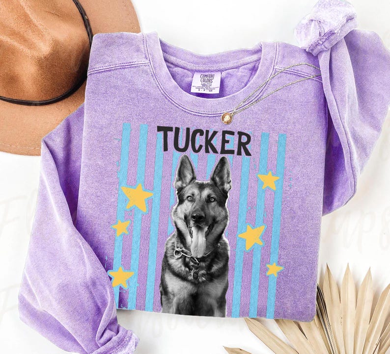 May include: A lilac-colored sweatshirt featuring the name "TUCKER" above a black and white image of a German Shepherd dog. The design includes blue vertical stripes and yellow stars. A gold necklace is draped across the sweatshirt.