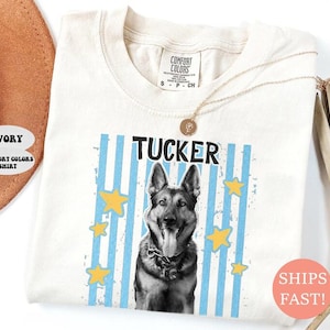 May include: Ivory Comfort Colors shirt featuring a German Shepherd dog graphic with the name "Tucker" in black text. The design includes blue vertical stripes and yellow stars. A gold necklace is draped over the shirt.