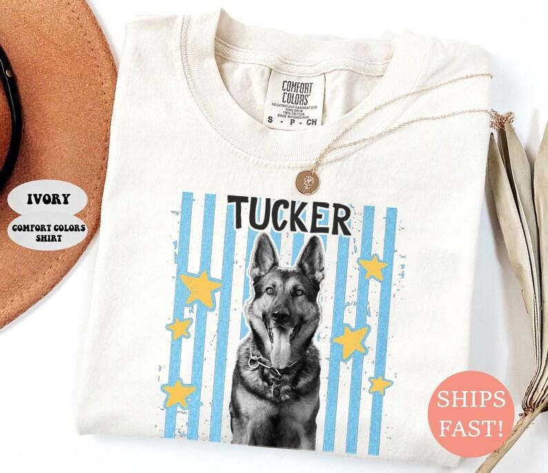 May include: Ivory Comfort Colors shirt featuring a German Shepherd dog graphic with the name "Tucker" in black text. The design includes blue vertical stripes and yellow stars. A gold necklace is draped over the shirt.