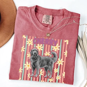 May include: A red Comfort Colors t-shirt featuring a dog graphic with the word "CHERRY" in purple. The design includes a dog illustration, yellow flowers, and vertical stripes in pink and teal.