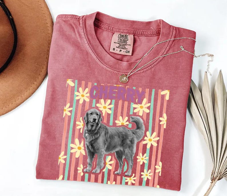 May include: A red Comfort Colors t-shirt featuring a dog graphic with the word "CHERRY" in purple. The design includes a dog illustration, yellow flowers, and vertical stripes in pink and teal.