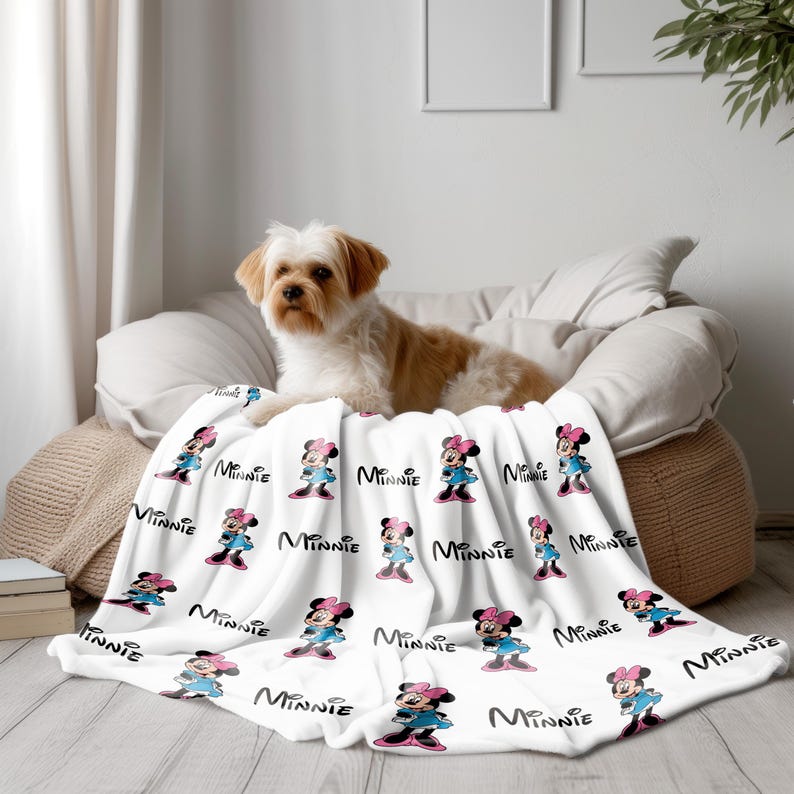 May include: A white blanket featuring a repeating pattern of Minnie Mouse in a blue dress and pink bow. The word "Minnie" is written in black script. A small dog rests on the blanket.