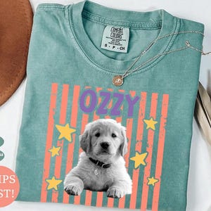 May include: Light green Comfort Colors t-shirt with the name "OZZY" in purple letters, a graphic of a golden retriever puppy, and yellow stars. The shirt has a crew neck and short sleeves. A silver necklace with a pendant is on the shirt.