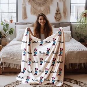 May include: A cream-colored blanket featuring a repeating pattern of Minnie Mouse in a blue dress and pink bow, with the name "Minnie" printed above each character. The blanket is displayed on a bed.