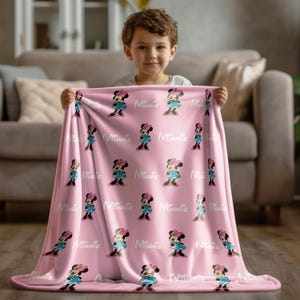 May include: A pink blanket featuring a repeating pattern of Minnie Mouse in a blue dress and pink bow. The word "Minnie" is printed in white between each character. The blanket is held by a child.