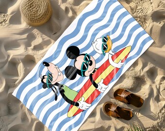 May include: Beach towel featuring Mickey Mouse, Donald Duck, and Goofy on a surfboard. The towel has a blue and white striped background. A straw hat and brown sandals are also visible on the sand.