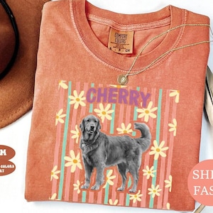 May include: Coral Comfort Colors t-shirt with a graphic of a Golden Retriever and the word "CHERRY" in purple. The design includes vertical stripes and daisy flowers. A gold necklace is draped on the shirt.