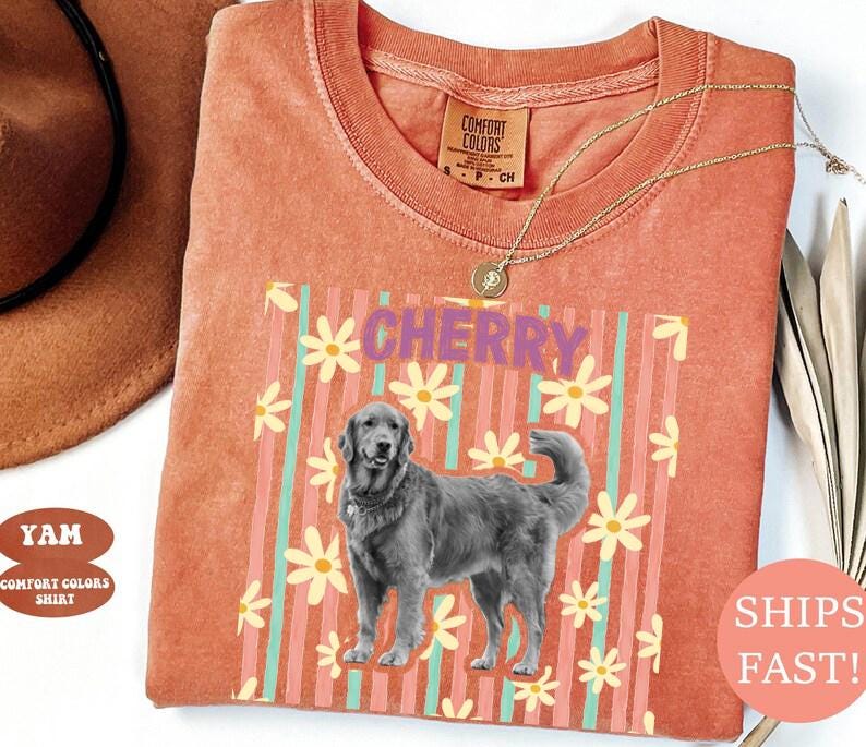 May include: Coral Comfort Colors t-shirt with a graphic of a Golden Retriever and the word "CHERRY" in purple. The design includes vertical stripes and daisy flowers. A gold necklace is draped on the shirt.