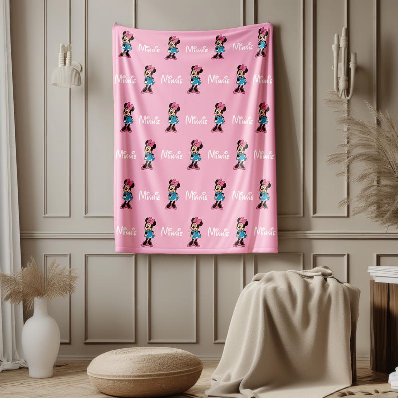May include: Pink blanket featuring a repeating pattern of Minnie Mouse in her classic outfit. The design includes Minnie's image and the word "Minnie" in white. The blanket is displayed against a neutral wall.