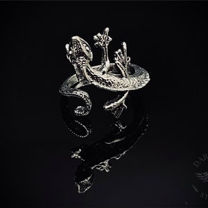 May include: A silver ring featuring a detailed gecko design. The gecko is depicted with its arms raised, as if in a celebratory gesture. The ring is set against a black background.