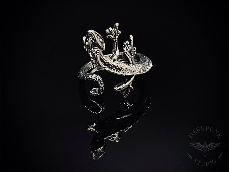 May include: A silver ring featuring a detailed gecko design. The gecko is depicted with its arms raised, as if in a celebratory gesture. The ring is set against a black background.