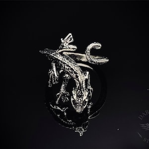 May include: A silver ring featuring a detailed lizard design with a curled tail. The lizard is positioned on top of the ring band, creating a three-dimensional effect.