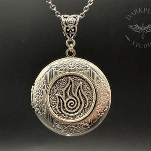 May include: Silver locket necklace with an intricate design featuring a stylised flame symbol. The locket has a hinged opening and a chain.
