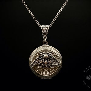 May include: A silver locket necklace with a detailed design of a moth with crescent moons on its wings. The locket is open and the inside is not visible.