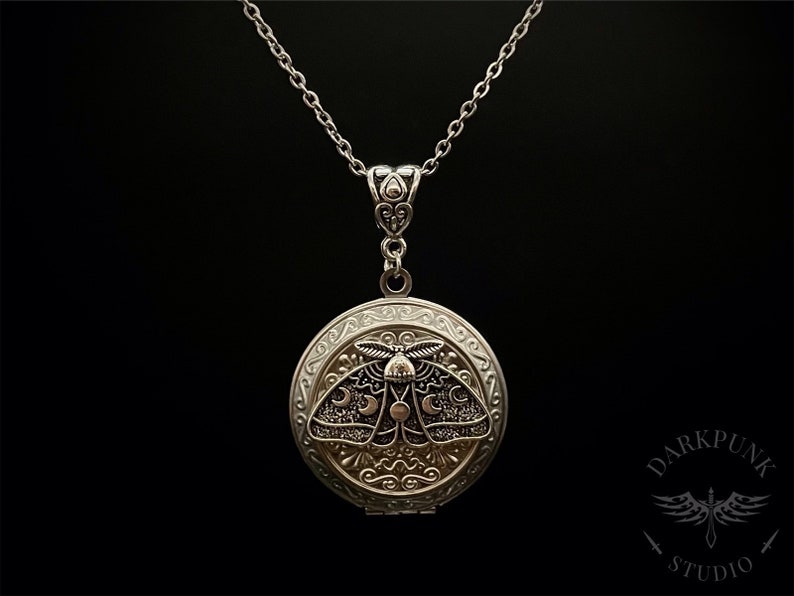 May include: A silver locket necklace with a detailed design of a moth with crescent moons on its wings. The locket is open and the inside is not visible.