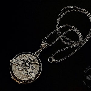 May include: A silver locket necklace with a detailed design of a moth and a heart. The chain is silver and has a lobster clasp.