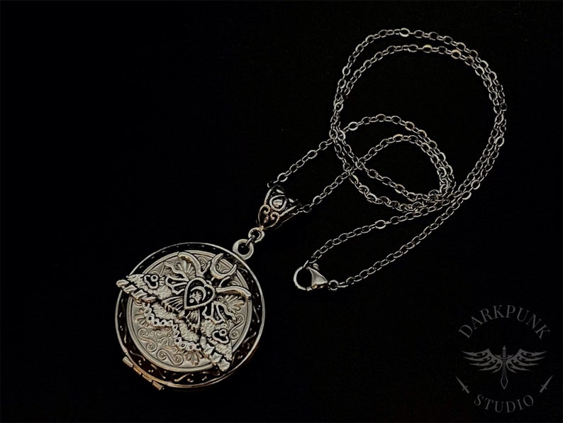 May include: A silver locket necklace with a detailed design of a moth and a heart. The chain is silver and has a lobster clasp.