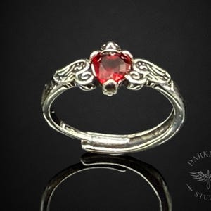 May include: A silver ring with a heart-shaped, deep red gemstone at the centre. The ring features ornate, swirling designs on the band and around the gemstone. The ring is set against a dark background.