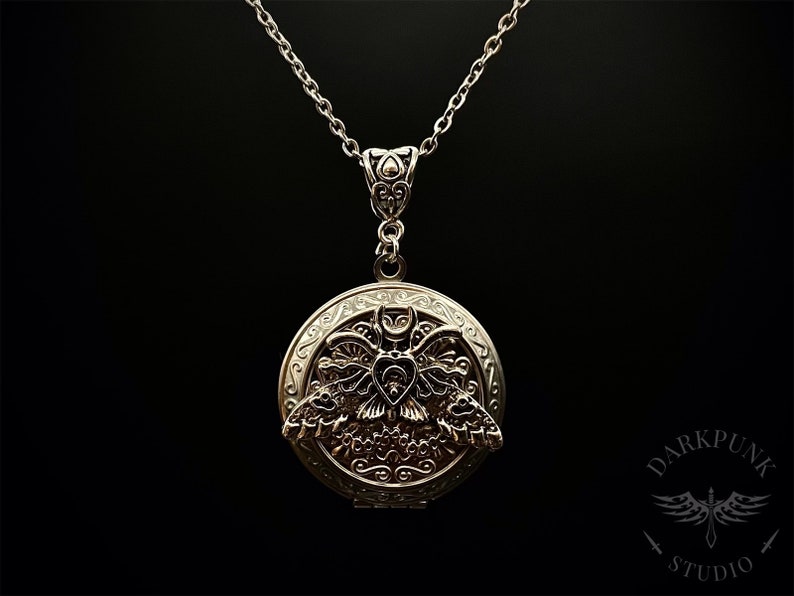 May include: A silver locket necklace with a detailed design of a moth with open wings. The locket is round and has a delicate chain.