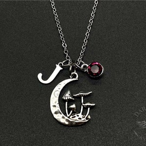 May include: A silver necklace with a crescent moon charm featuring three mushroom silhouettes. The necklace also includes a letter 'J' charm and a dark red gemstone.