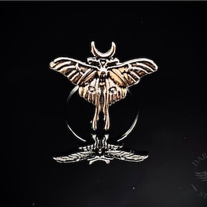 May include: A silver ring featuring a detailed, three-dimensional design of a moth with a crescent moon on its head.