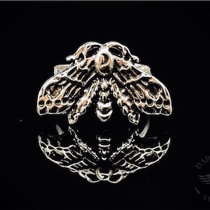 May include: A silver ring featuring a detailed, three-dimensional design of a moth with intricate wings and a skull-like head.
