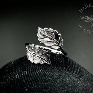 May include: Silver ring featuring two leaf designs, one wrapped around the other. The ring is displayed on a black fabric surface.