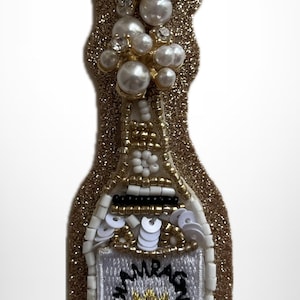 May include: A champagne bottle-shaped patch featuring a gold glitter background, white and gold seed beads, white sequins, and a cluster of white faux pearls and clear crystals at the top. A white label reads CHAMPAGNE with a small gold embroidered crown.