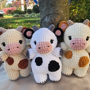 Könnte beinhalten: Drei gehäkelte Plüsch-Kuhspielzeuge. Eines ist weiß mit schwarzen Flecken, eines ist cremefarben mit braunen Flecken und eines ist cremefarben mit orangefarbenen Flecken.