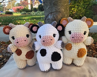 Puede incluir: Tres juguetes de peluche de vaca hechos a crochet. Uno es blanco con manchas negras, otro es crema con manchas marrones y el último es crema con manchas naranjas.