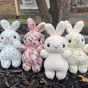 Puede incluir: Cuatro peluches de conejo de crochet en diferentes colores. Los conejos están de pie sobre una cama de mantillo marrón. Los conejos tienen todos la misma expresión, con ojos grandes y una nariz pequeña.