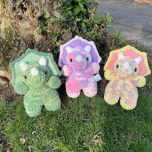 May include: Three crocheted plush triceratops dinosaurs in different colors: green, pink and yellow. The dinosaurs are sitting on a bed of green grass.