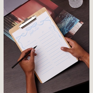 May include: A clipboard with a white paper pad and a blue ribbon design. The paper has the words "Gift List" written at the top and a list of blank lines with checkboxes next to each line. A person is writing on the clipboard with a black pencil.