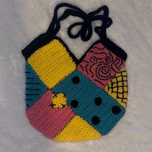 Peut inclure: Un sac fourre-tout crocheté à la main avec un motif patchwork jaune, rose et turquoise. Le sac a des accents noirs, y compris les sangles et les bords du patchwork. Le design comprend des points noirs et un patch en forme de fleur.
