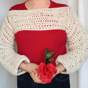 May include: A white crocheted sweater with long sleeves and a wide, open neckline. The sweater is worn over a red shirt. The person is holding a red rose.